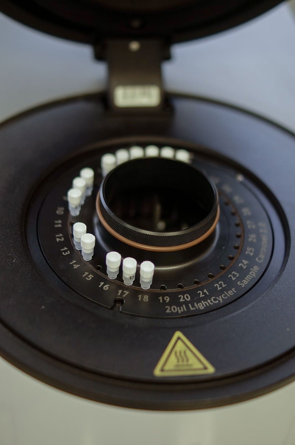 Close-up of a laboratory centrifuge rotor holding multiple small white sample tubes arranged in a circular pattern, with a warning symbol visible in the lower part of the image.