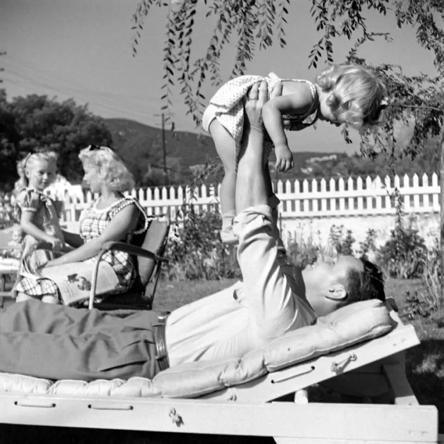A man lies on a lounge chair, lifting a smiling young girl above him. In the background, two women and another girl sit and talk near a white picket fence in a sunny yard.