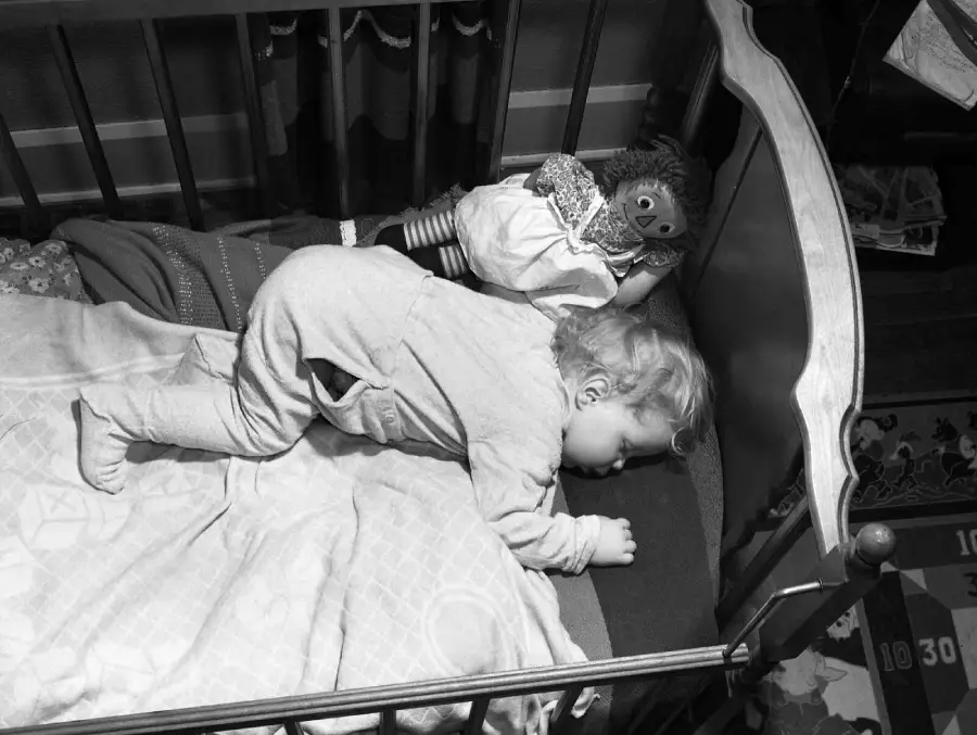 A small child sleeps face down in a crib, wearing pajamas. A rag doll is propped up near the child, and a blanket covers part of the bed. The scene is calm and peaceful.