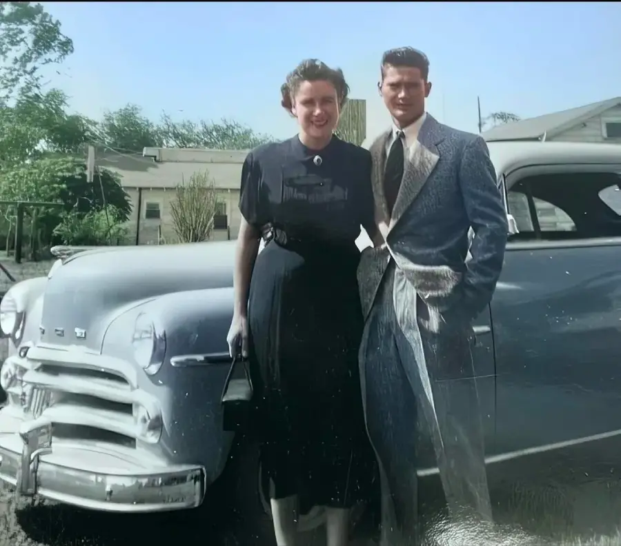A woman in a dark dress and a man in a suit stand smiling beside a vintage car, possibly from the 1940s or 1950s, with a house and trees in the background.