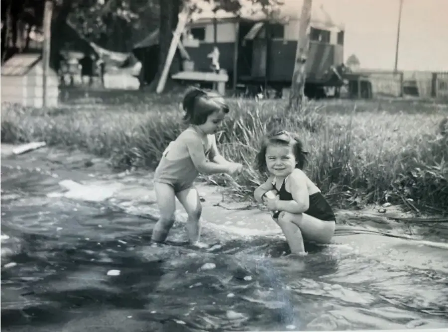 Two young girls play and smile in shallow water near a grassy shore. In the background are trees, a fence, and a trailer or cabin, giving a vintage, outdoor, summer atmosphere.