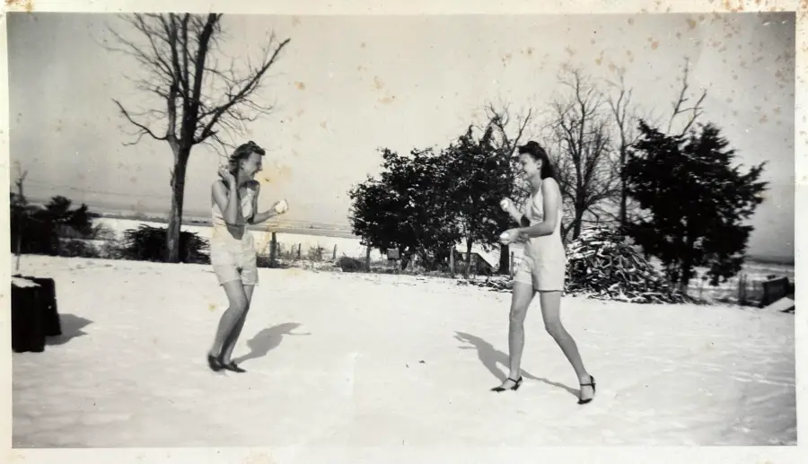 Two women wearing shorts and tank tops playfully face off in a boxing stance outside on a snowy field, with trees and bushes in the background.