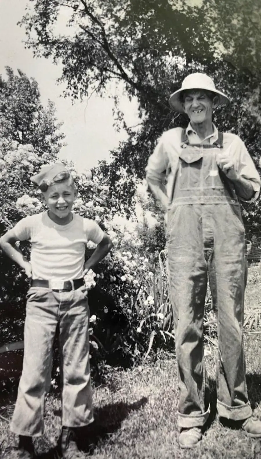 A smiling boy and older man stand outside in work clothes and hats, surrounded by trees and flowers, posing for a black-and-white photo.