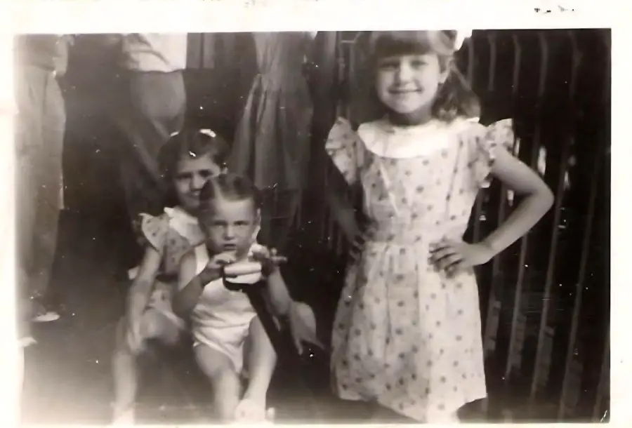 Three young children pose for a photo; one girl stands in front smiling with a hand on her hip, while two other children sit close together behind her. The image is black and white.
