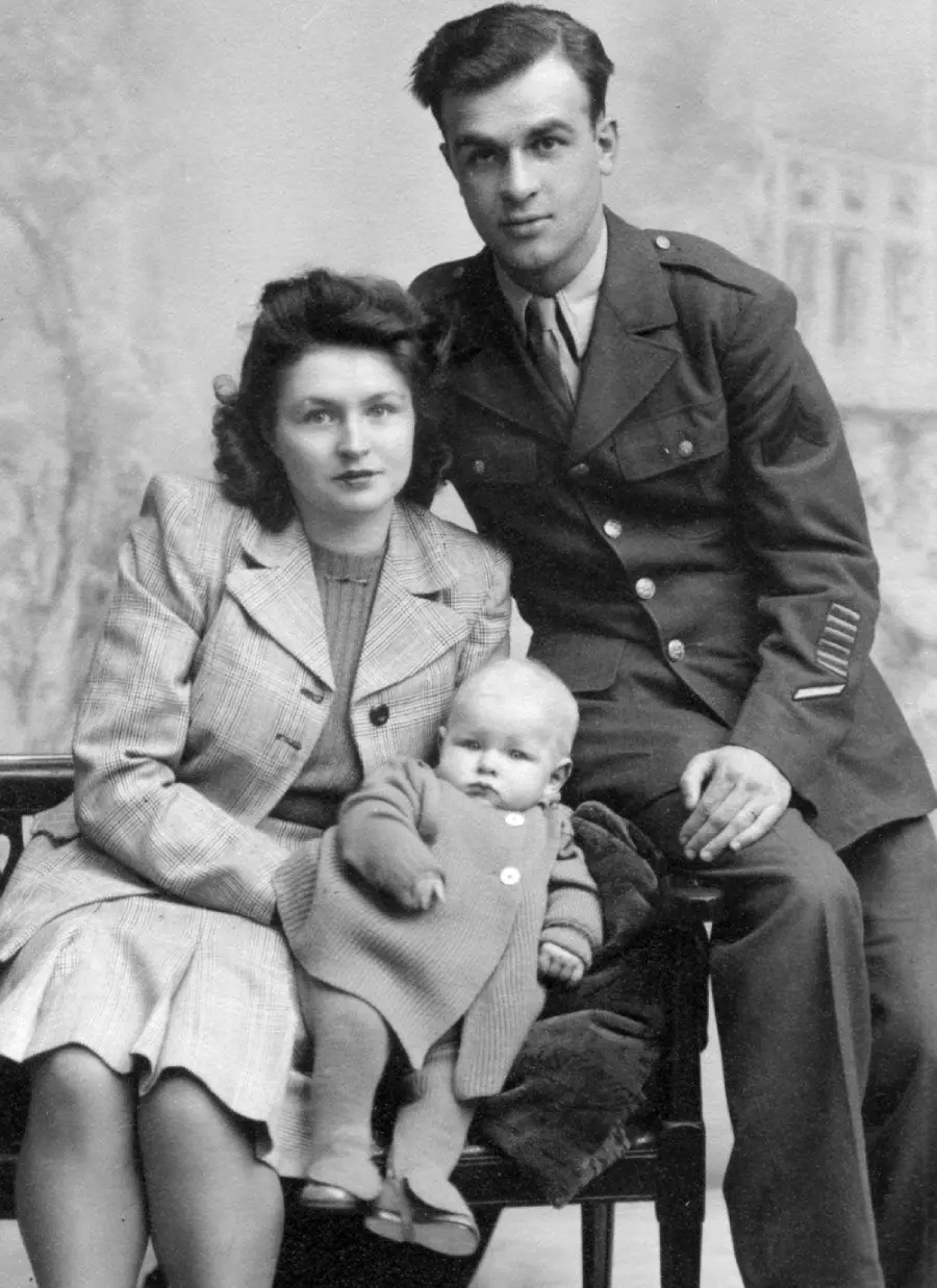 A black-and-white photo of a woman in a coat, a man in a military uniform, and a baby in a sweater sitting on a bench, posed for a formal portrait with a vintage backdrop.