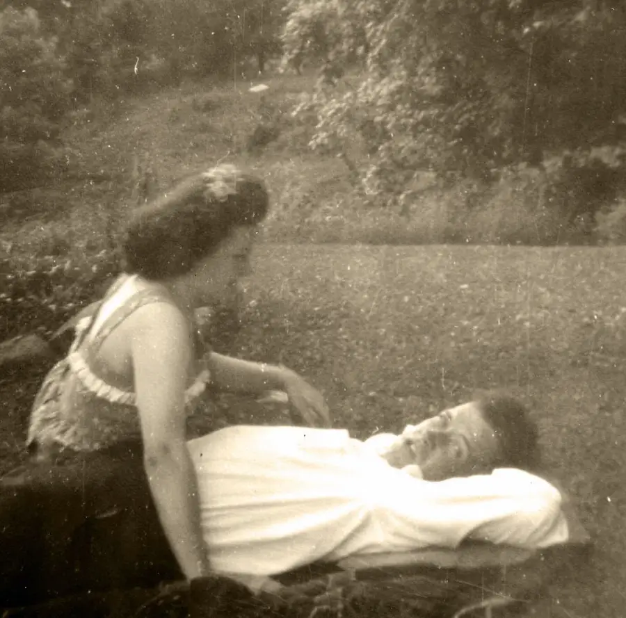 A woman in a dress leans over a man lying on a blanket outdoors, with trees and grass in the background. The image is in black and white and appears vintage.