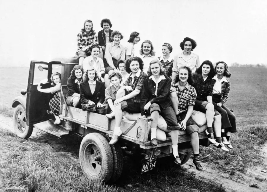 A group of young women sit and pose happily on the back of a truck in a field, wearing casual clothing, likely from the mid-20th century. The setting appears rural with grass and open land in the background.