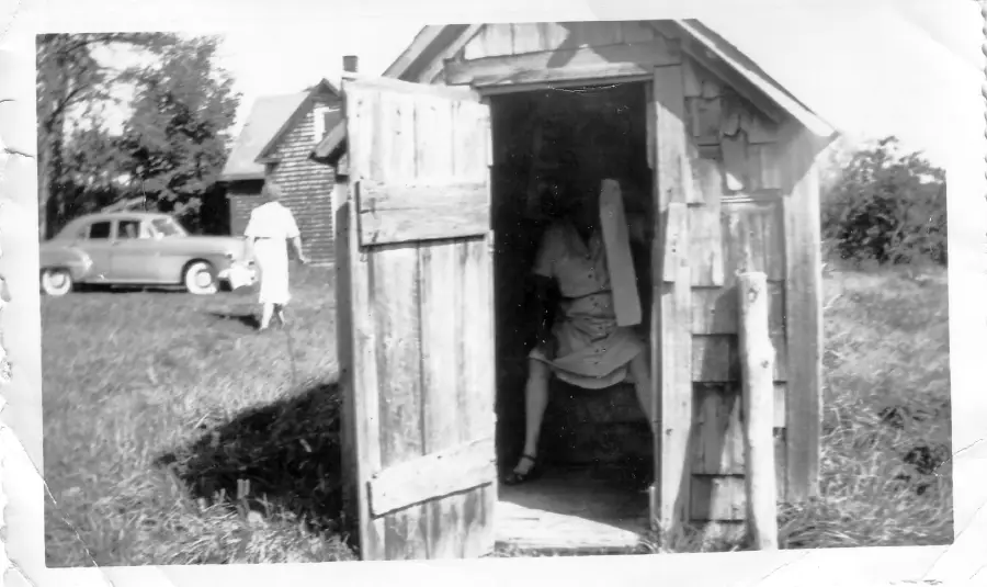 A woman sits inside a small wooden outhouse with the door open. There is a vintage car and a person walking in the grassy yard, with another small building in the background.