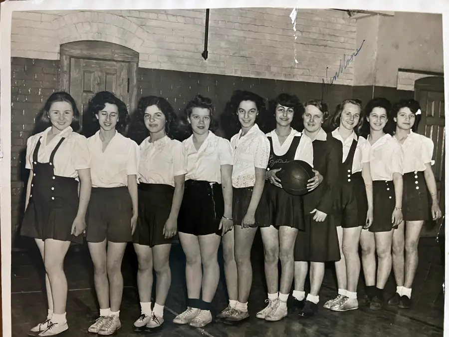 A vintage black-and-white photo of ten young women and one older woman, all in athletic attire, posing indoors. The woman in the center holds a basketball. They stand in a row against a brick wall.
