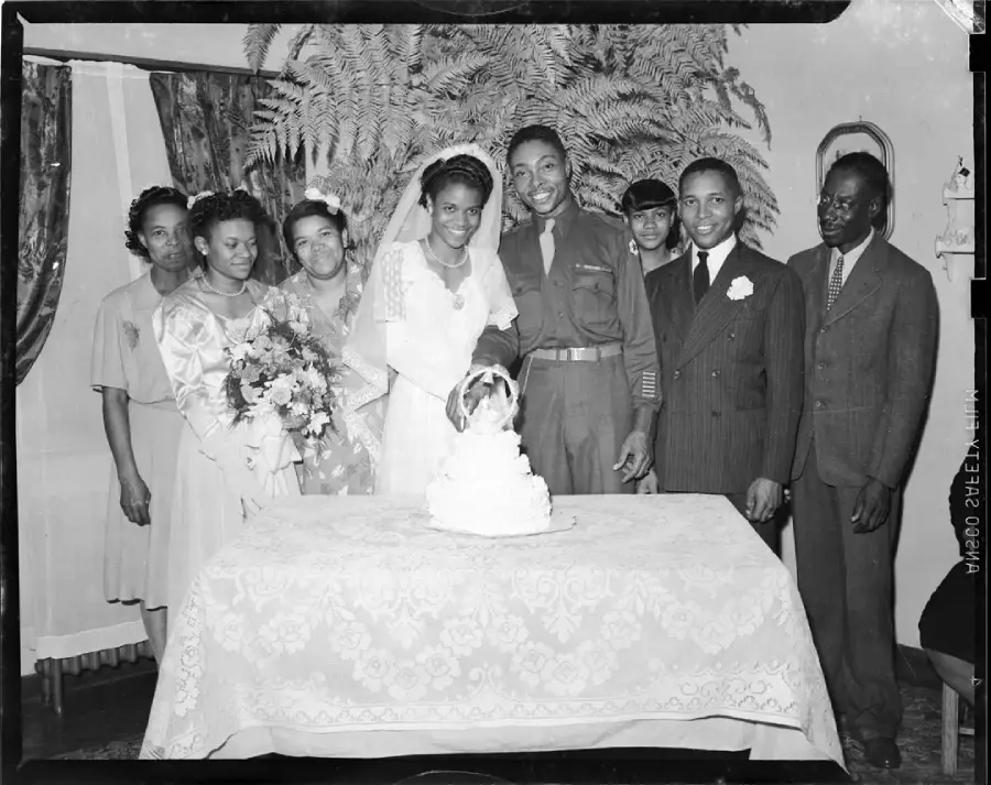 A bride and groom stand behind a cake with family and friends, all smiling, in a formal setting with a lace-covered table and floral decorations.