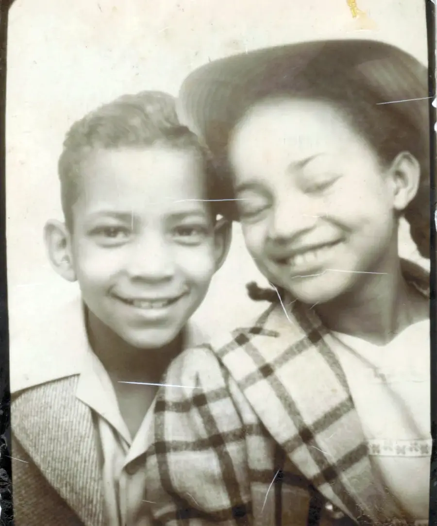 A vintage black-and-white photo of two children smiling. The boy on the left wears a light jacket, while the girl on the right, with her eyes closed, wears a plaid coat and a hat. Both look happy and close together.