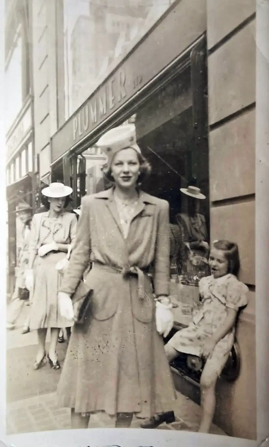 Vintage photo of a woman in a dress and hat standing on a city sidewalk near a shop called “PLUMMER,” with two women and a young girl sitting nearby. All wear 1940s-style clothing.