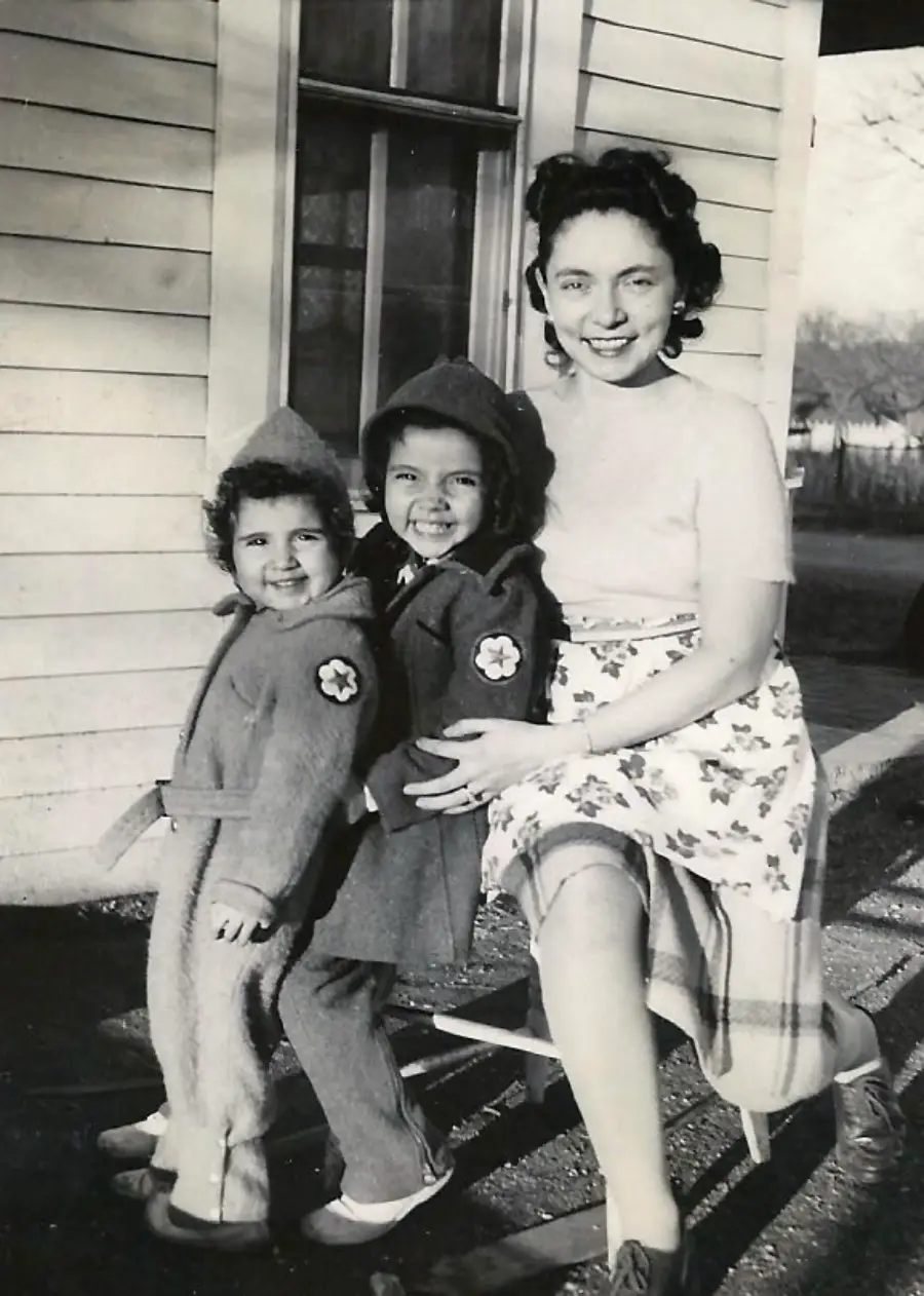 A woman sits on a bench outside with two young children in coats and hoods, all smiling at the camera. The scene is in front of a wooden building, with trees visible in the background.