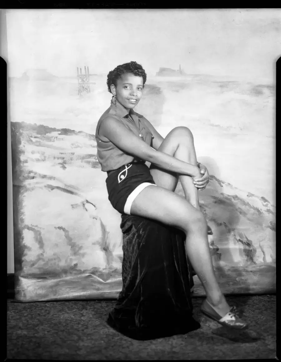 A woman sits on a stool with her legs crossed, smiling at the camera. She wears a sleeveless top, shorts, hoop earrings, and loafers. The backdrop shows a scenic, painted landscape.
