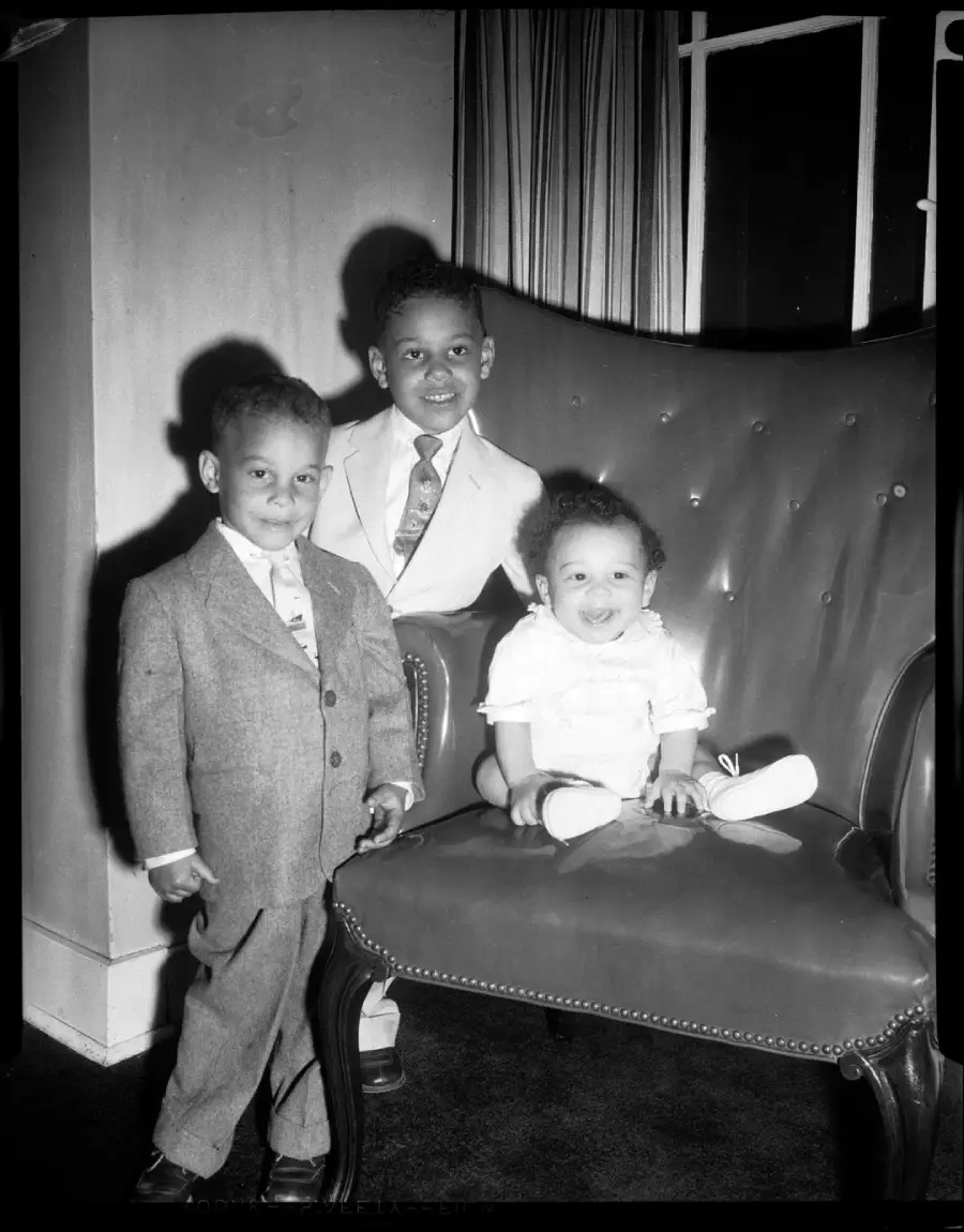 Three young children pose indoors. Two boys in suits stand beside a large armchair, smiling, while a baby in white sits on the chair, also smiling. A window with curtains is in the background.