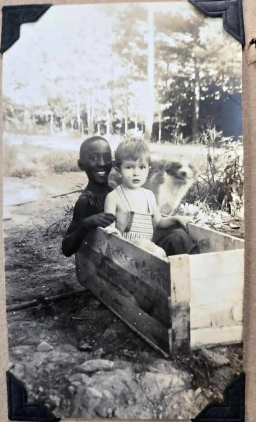 Two children sit in a wooden crate outdoors, smiling at the camera. A dog is partially visible behind them. The photo appears old and is in black and white.