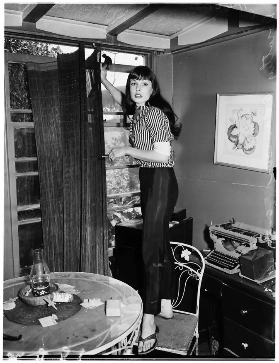 A woman in striped shirt and pants stands barefoot on a chair, adjusting a curtain in a room with a table, papers, typewriter, and framed picture on the wall.