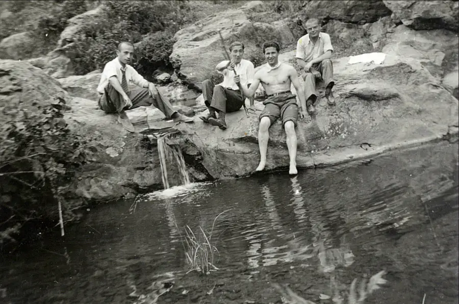 Four men relax by a rocky stream, with two sitting on rocks and two dipping their feet in the water. One man is shirtless. The setting appears natural and outdoors, surrounded by rocks and vegetation.