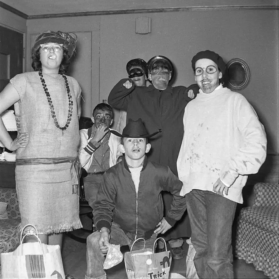 Five people in costume pose indoors with Halloween treat bags. Some wear hats, masks, or makeup. The group stands and kneels in a living room with a couch and chairs in the background. The photo is black and white.