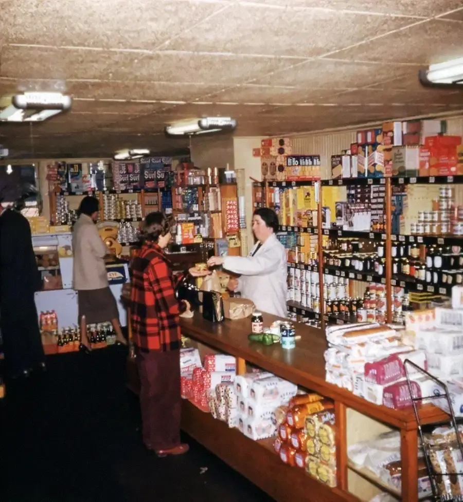 A grocery store with shelves filled with various products. A shopkeeper serves two customers at the counter, while another shopper browses in the background. The store appears organized and well-stocked.