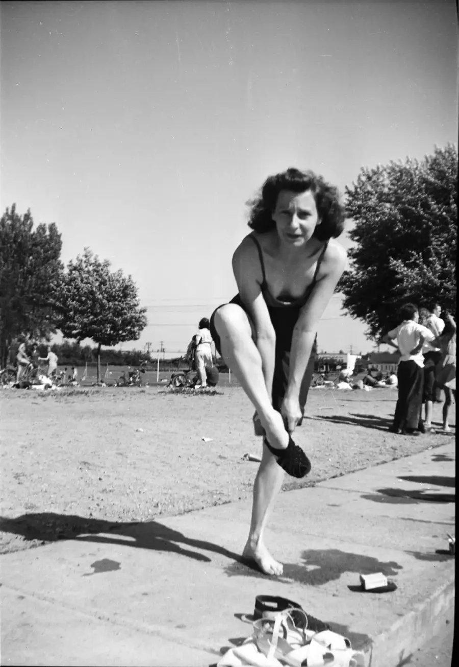 A woman in a swimsuit stands on a sidewalk at a beach or poolside, bending to put on a shoe. People and trees are visible in the background under a clear sky.
