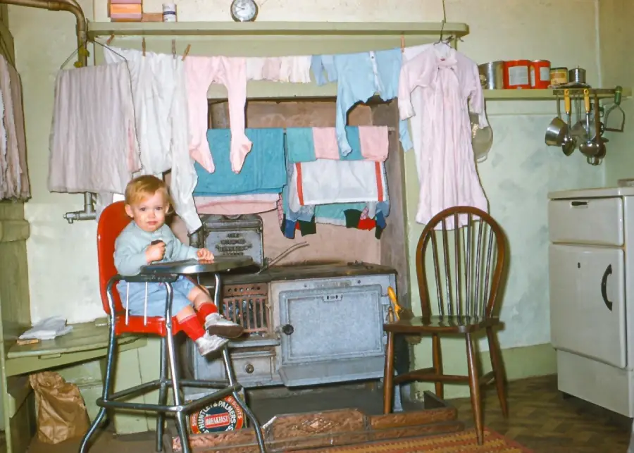 A toddler in a high chair sits in a vintage kitchen. Clothes hang to dry above an old stove, with pots, pans, and kitchen items nearby. The room has a warm, nostalgic feel.