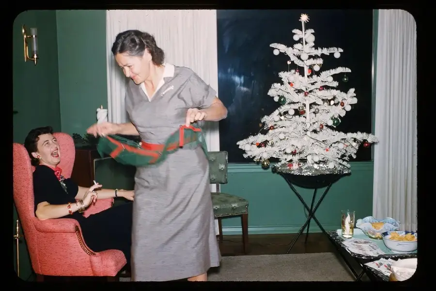 Two women enjoy a festive moment in a living room with a white Christmas tree. One woman sits laughing in a pink chair, while the other stands holding a red and green apron. Snacks and drinks are on a table nearby.
