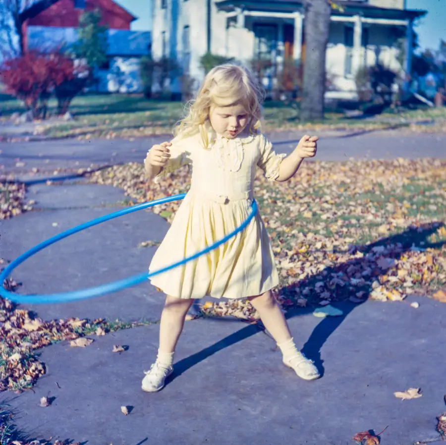 A young girl in a yellow dress plays with a blue hula hoop outside on a sidewalk, with autumn leaves scattered around and houses in the background.