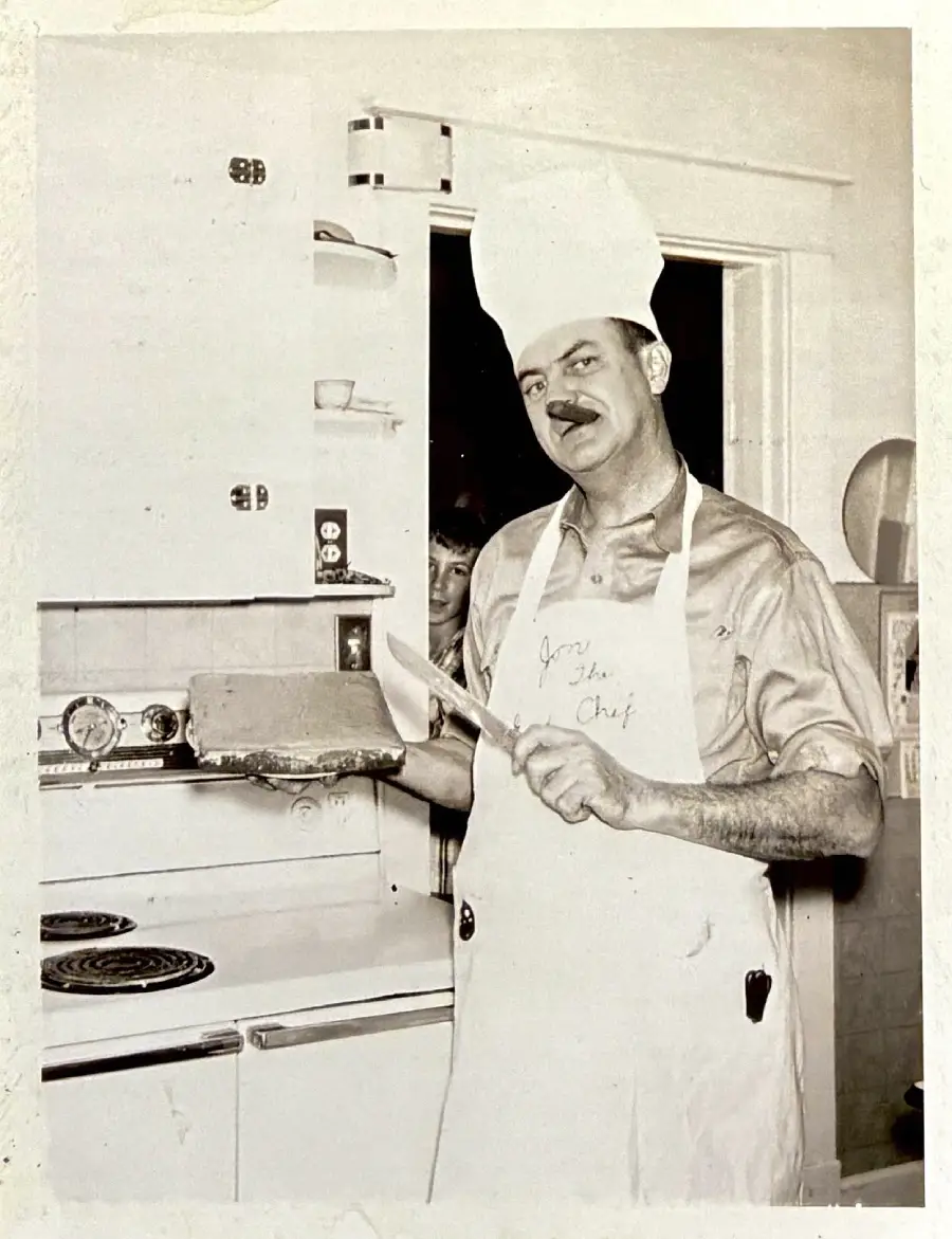 A man in a chef’s hat and apron smiles while holding a cake and a knife in a kitchen. A person peeks from behind a door in the background. The photo appears to be vintage and in black and white.