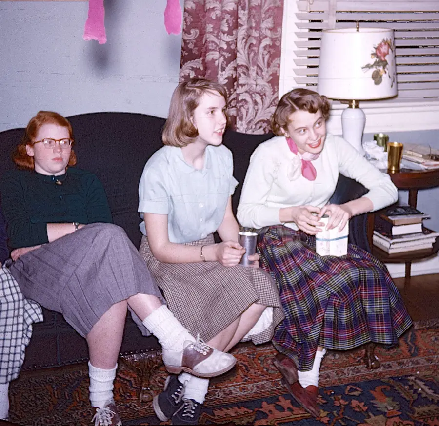 Three young women in vintage clothing sit on a sofa. Two are smiling and engaged, while one looks serious. A lamp, books, and a window with blinds are in the background. Pink streamers hang from above.