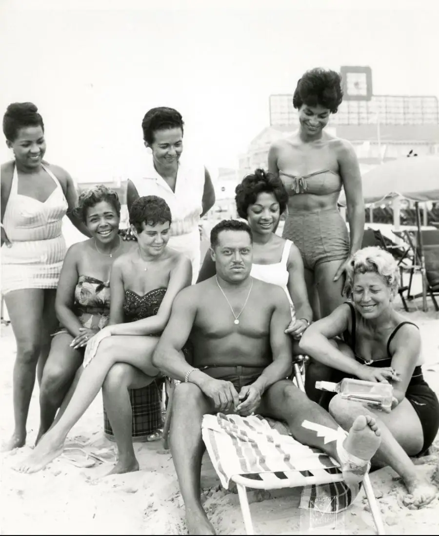 Seven women and one man in vintage swimsuits pose together on a sandy beach; most are smiling, and the man sits on a chair in front with women seated and standing around him. A building is visible in the background.