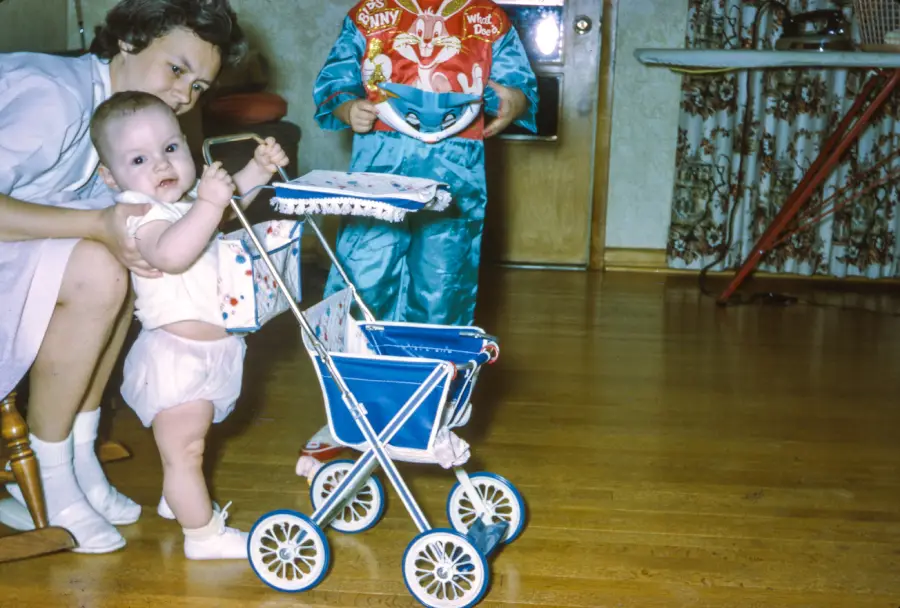 A baby stands holding a toy stroller with the help of an adult, while another child dressed in a colorful outfit stands nearby on a wooden floor. An ironing board and patterned curtains are visible in the background.