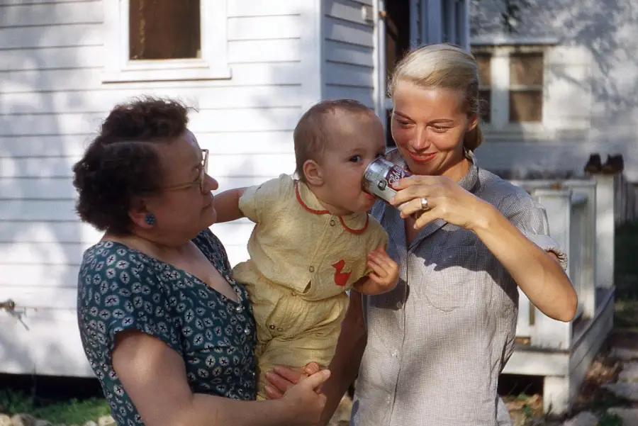 A woman holds a baby while another woman helps the baby drink from a can outside a white house. All three appear happy, enjoying a sunny day.