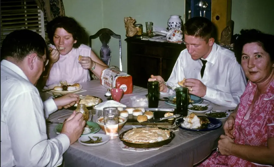 Four people sit around a dinner table eating a meal with pie, bread, green beans, and drinks. They appear to be enjoying a homemade dinner in a cozy dining room setting.