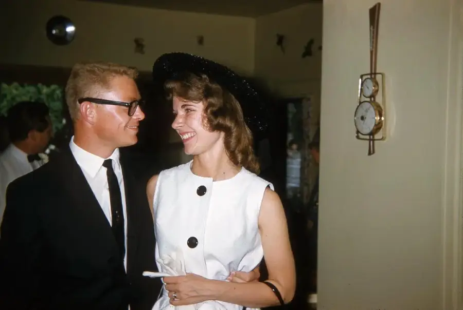 A man in glasses and a dark suit smiles at a woman in a white dress and black hat. They appear happy, standing indoors near a vintage wall clock, with other people in the background.