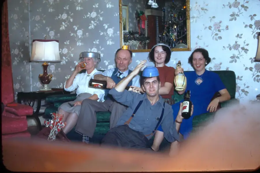 Five people sit in a living room, wearing pots and pans as hats, holding bottles and smiling. There’s a lamp, floral wallpaper, and a mirror reflecting decorations in the background.