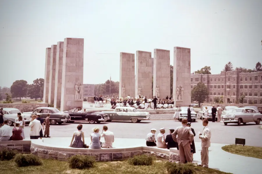 A crowd gathers near a monument with tall stone pillars and statues, as a band performs. Vintage cars are parked nearby, with people sitting and standing around on a sunny day.