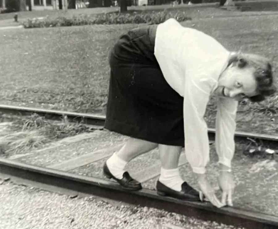A woman in a skirt and blouse bends over and touches her toes while standing on a railroad track, looking back at the camera. The photo is in black and white.