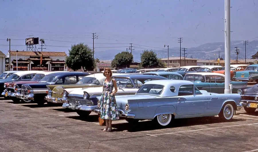 A woman in a floral dress stands next to a light blue vintage car in a sunny parking lot filled with classic cars from the 1950s, with buildings and mountains visible in the background.