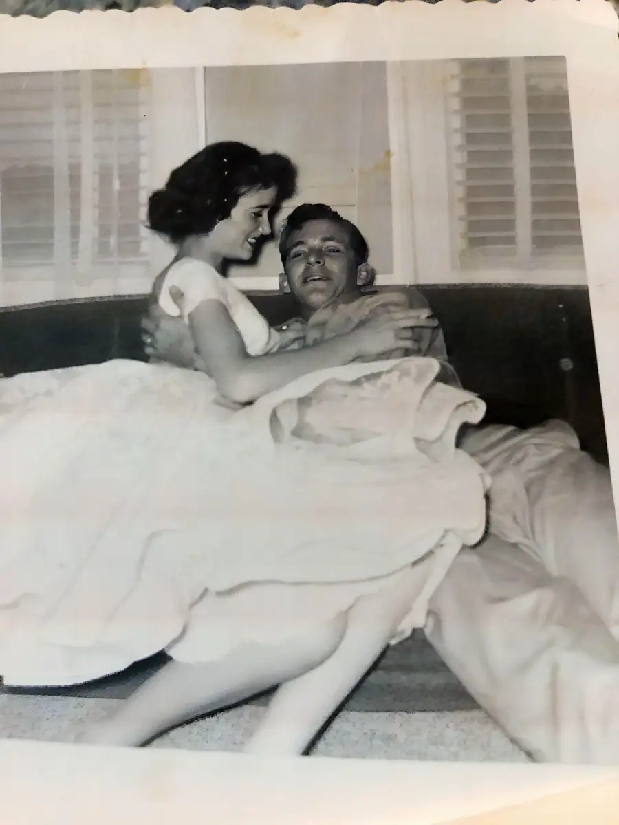 A smiling woman in a dress sits on a man’s lap as they relax together on a couch in a black and white photo. The man lounges back, holding the woman, and both appear happy.