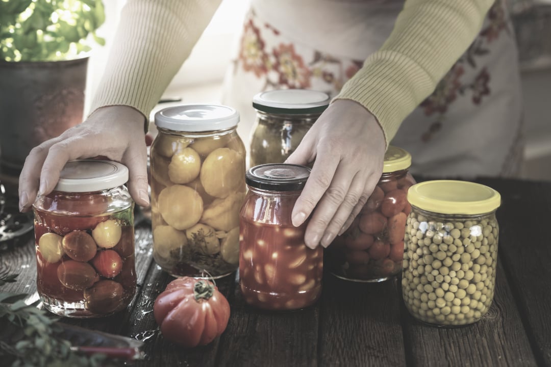A person arranges jars of preserved vegetables, including tomatoes and beans, on a wooden table. Sunlight shines through a window in the background.