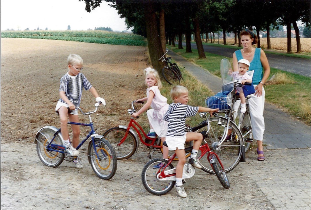 A woman stands on a path with a baby in her arms, while three children on bikes and one child on a tricycle are nearby. Trees and fields are in the background.