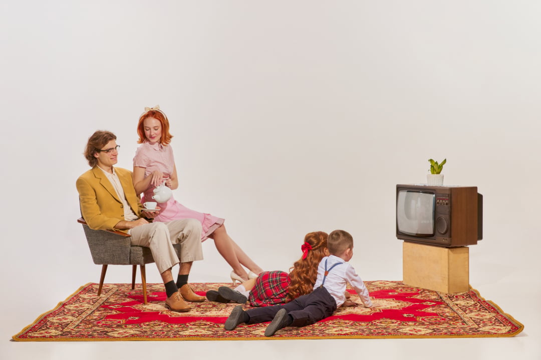 A man and woman sit on a chair and the floor with tea, while two children lie on a red carpet watching an old TV on a box. The scene is styled with vintage decor.