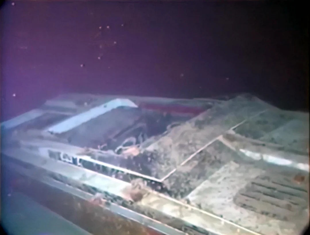 An underwater image shows part of a large, decaying shipwreck resting on the ocean floor, with corroded metal, sediment buildup, and dim lighting.