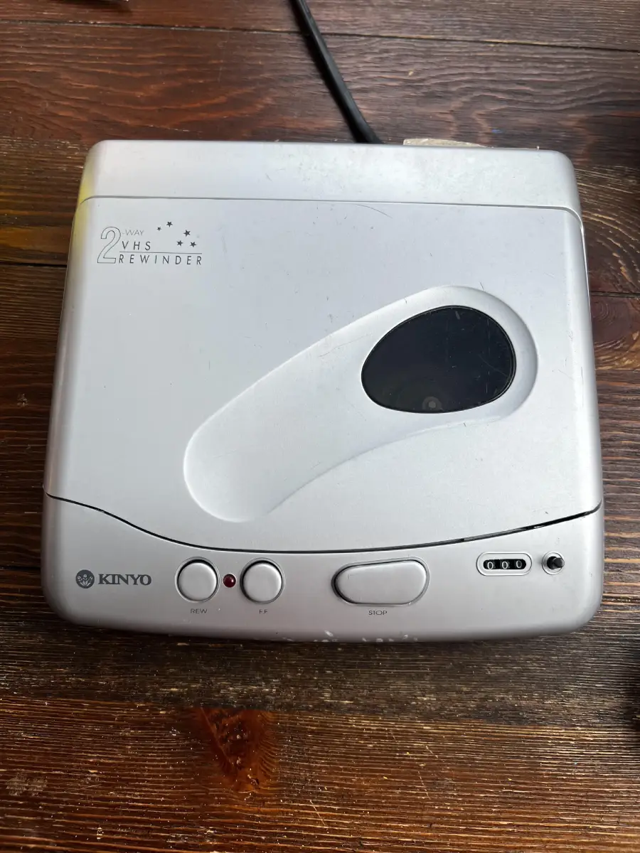 A silver Kinyo VHS tape rewinder with control buttons sits on a wooden surface. A black power cord extends from the top of the device.
