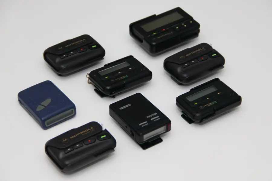 Eight vintage pagers of various brands and styles are arranged on a white surface, showing buttons and small screens, with most pagers being black and one blue.