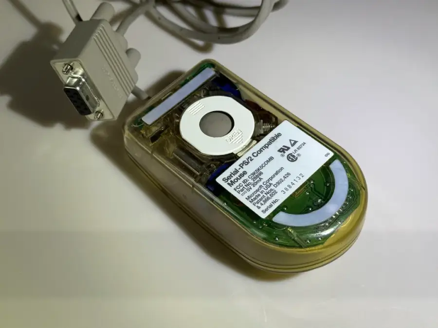 A translucent computer mouse with a visible circuit board, a round white trackball, and a serial port connector, placed on a white surface.