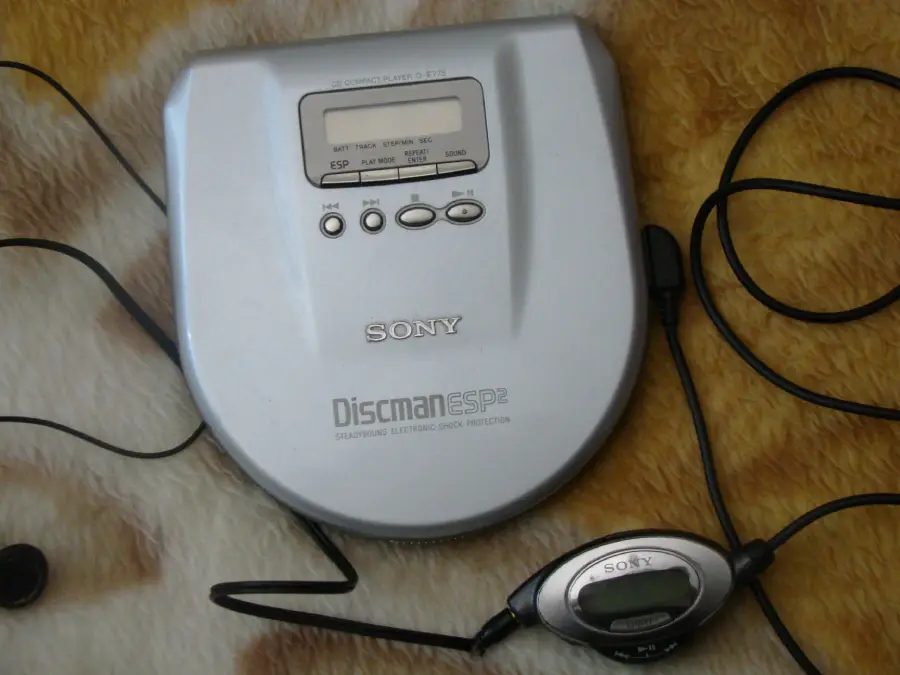 A silver Sony Discman ESP2 portable CD player with attached black wired headphones, resting on a fuzzy brown and beige surface. The player’s display screen and control buttons are visible.