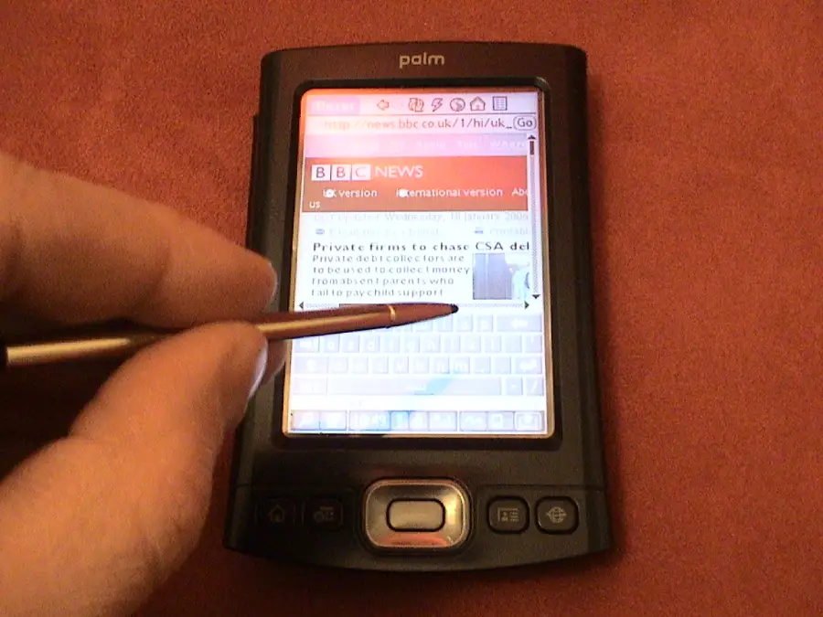 A hand holding a stylus is interacting with a Palm handheld device displaying a BBC News webpage on its touchscreen with a virtual keyboard visible.