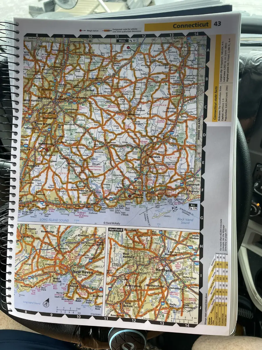 A spiral-bound road atlas is open to a detailed map of Connecticut, showing highways, cities, and insets of New Haven and Hartford. The book rests on a car’s steering wheel.